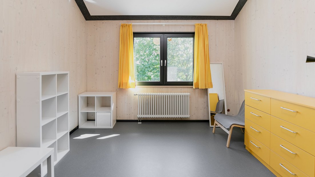 Habitación en el Rosenhaus de arriba con cómoda amarilla, estantes blancos, mesa, espejo y sillón gris